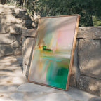 May include: Abstract art print in a wooden frame. The artwork features a blend of colors, including pink, green, yellow, and beige, creating a soft, flowing composition. The painting is leaning against a stone wall.
