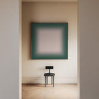May include: A square wall art piece featuring a dark green border that fades into a light beige center, displayed above a small black stool with a circular seat and a rectangular backrest, positioned in a room with light-colored walls.