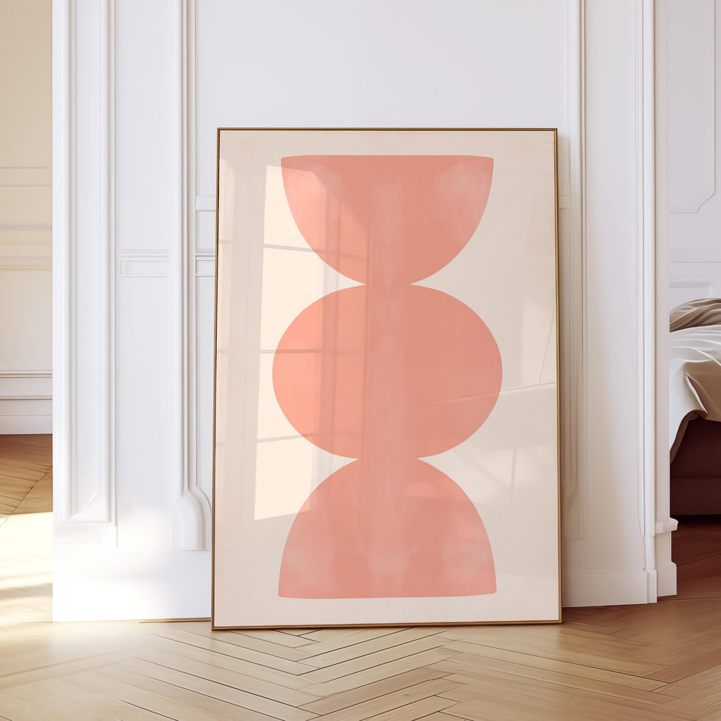 May include: A rectangular framed wall art print featuring a vertical arrangement of three stacked, soft coral-colored geometric shapes, including two semi-circles at the top and bottom and a central oval, set against a solid light beige background.