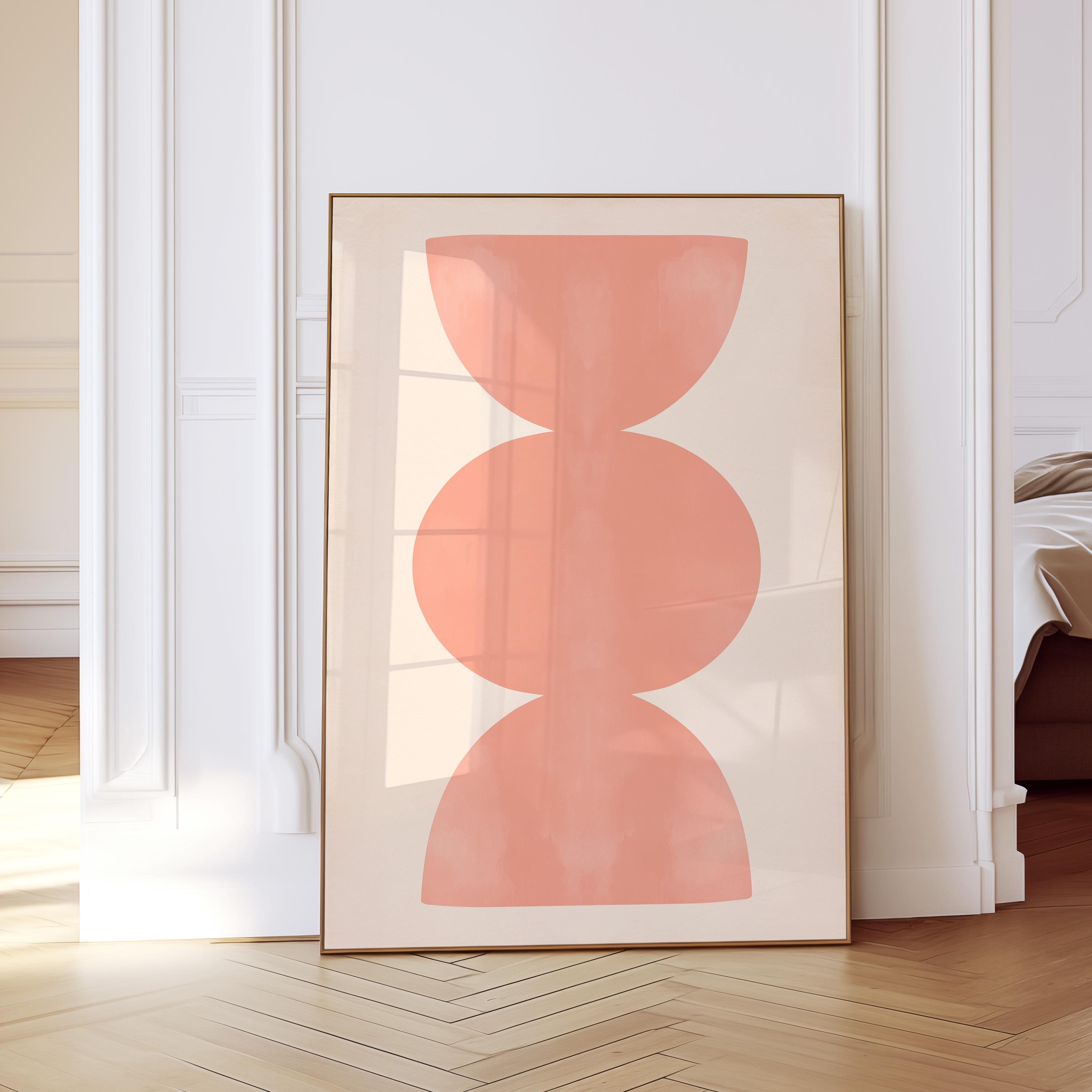 May include: A rectangular framed wall art print featuring a vertical arrangement of three stacked, soft coral-colored geometric shapes, including two semi-circles at the top and bottom and a central oval, set against a solid light beige background.