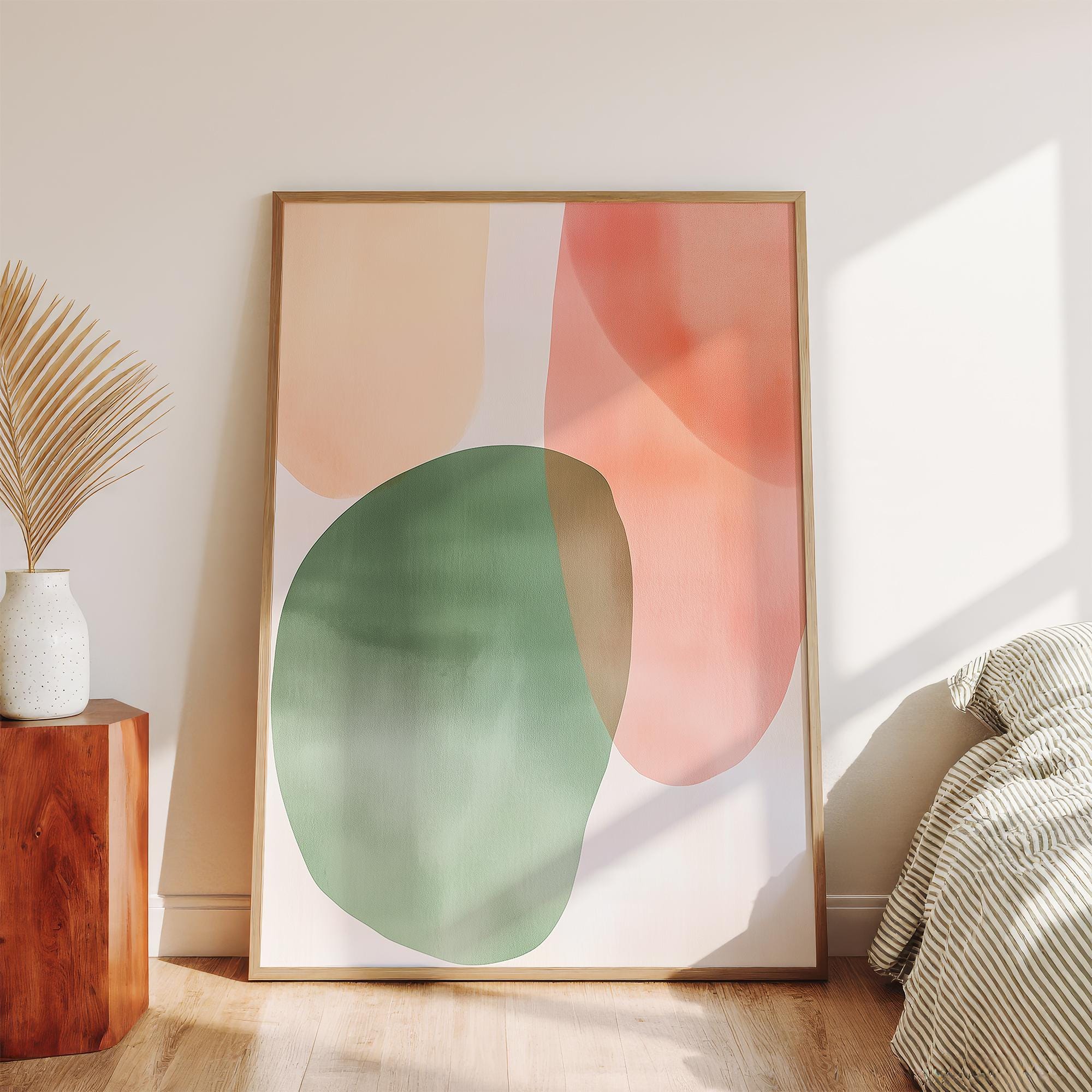 May include: Abstract art print featuring overlapping watercolor shapes in peach, green, and coral hues. The artwork is framed in a light wood frame and leans against a white wall. A minimalist aesthetic for home decor.