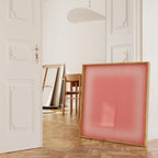 May include: A large, square artwork with a light pink translucent panel is set in a natural wood frame. The artwork leans against a white wall in a room with a wooden floor. Other artworks and a wooden chair are also visible.