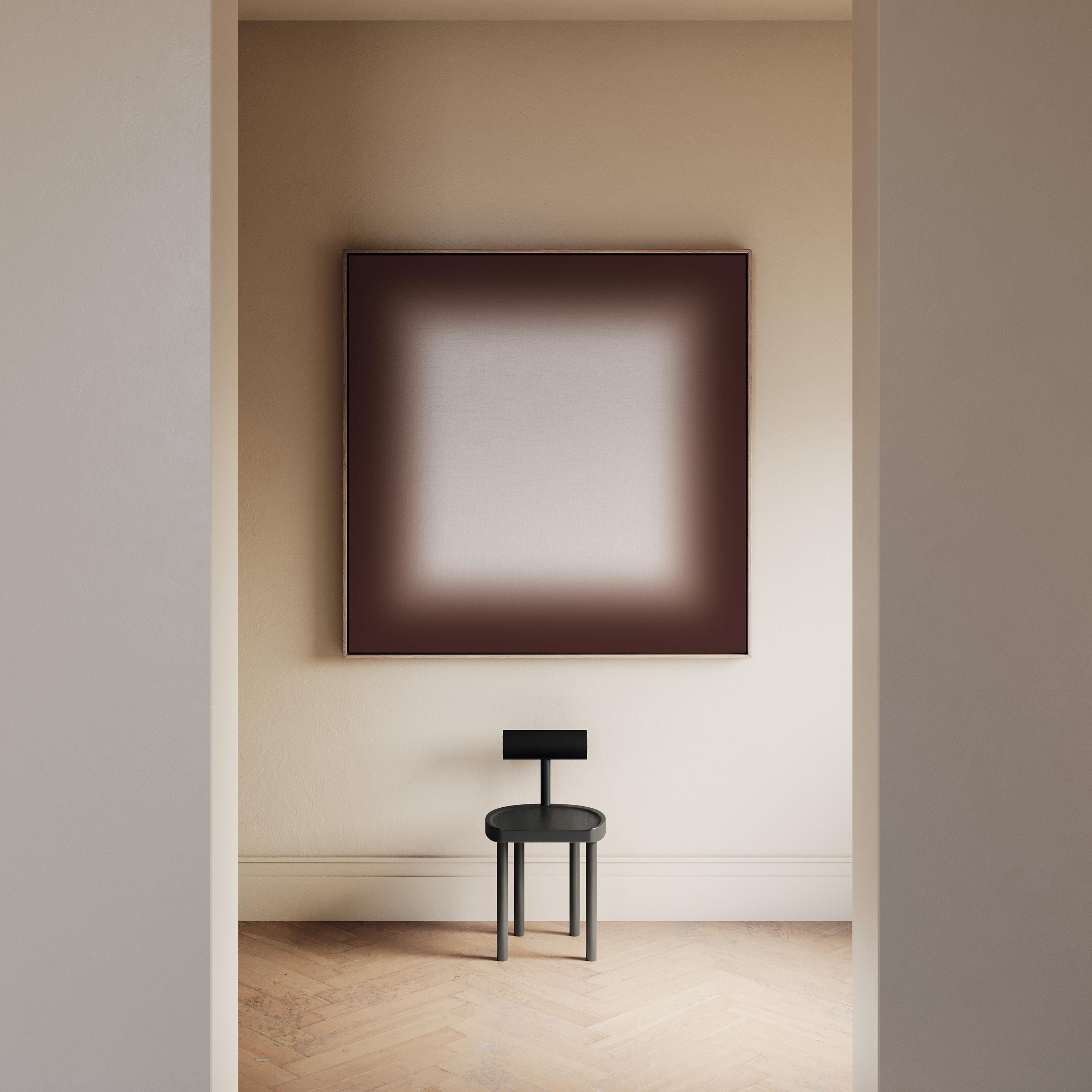 May include: A modern interior scene featuring a dark gray chair with a small backrest and a large framed artwork. The artwork has a gradient effect, transitioning from a dark brown border to a lighter center. The room has light-colored walls and a wooden floor.