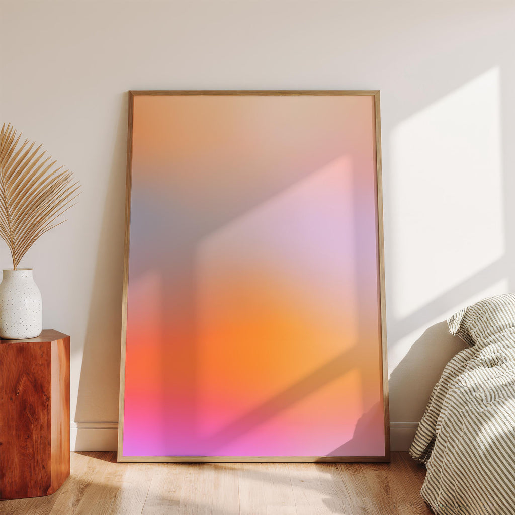 May include: A framed abstract art print with a gradient of warm colors. The artwork transitions from pink at the bottom to orange and then to a light blue-gray at the top. The print is displayed in a room with a wooden floor, a white wall, and a wooden side table with a vase and decorative plant.