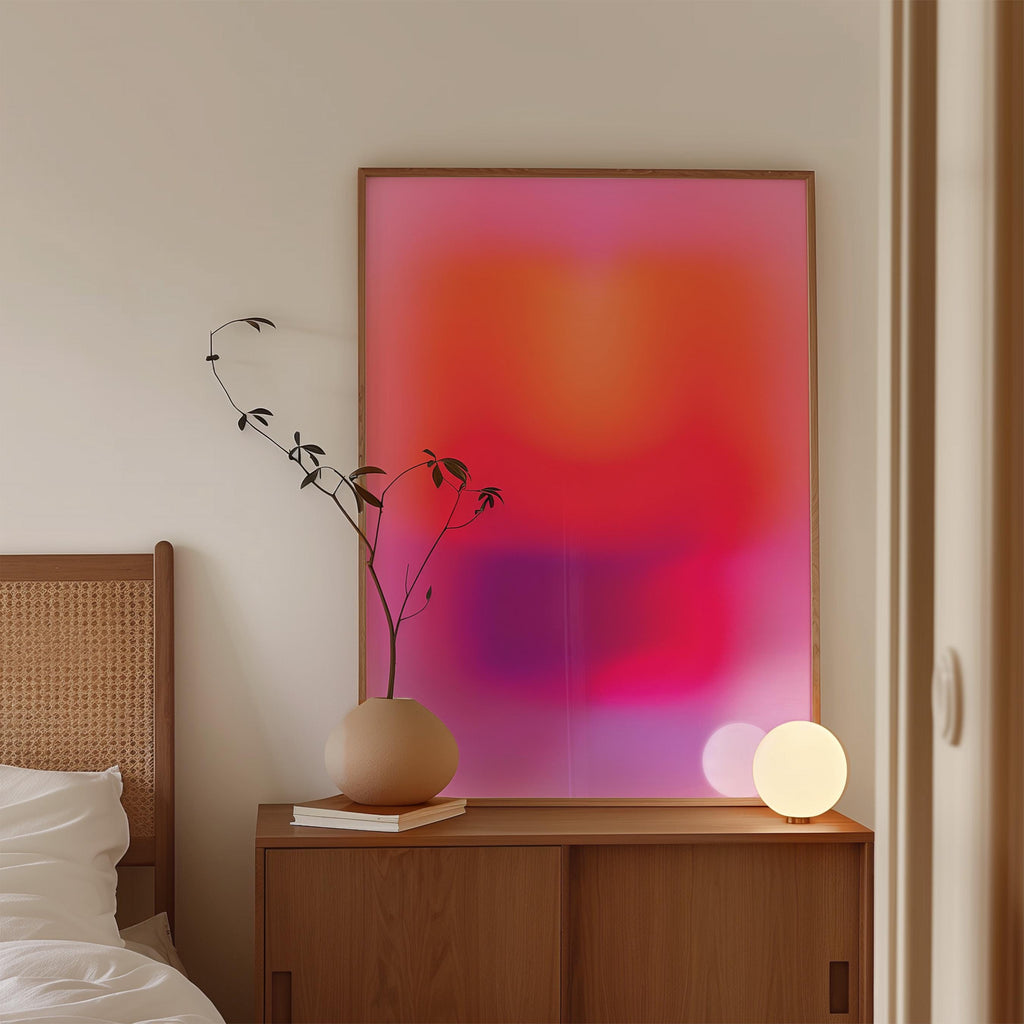 May include: A large framed abstract art print with a gradient of pink, orange, and purple hues. A wooden cabinet holds a beige vase with a sprig of greenery, a stack of books, and a glowing spherical lamp. A bed frame is visible on the left.