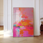 May include: Abstract art print with a vibrant color palette. The artwork features a blend of pink, orange, yellow, and blue hues. The painting is framed in a thin, gold-colored frame and leans against a white wall.
