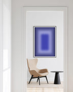 May include: A framed abstract art print with a gradient of blue hues, from dark to light, hangs on a white wall. A beige armchair and a small black side table are in the foreground, creating a minimalist interior design.