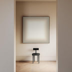 May include: A minimalist interior scene featuring a large, square artwork with a soft, gradient color scheme. Below, a small, dark gray chair with a circular seat and a rectangular backrest stands on a light-colored wooden floor.