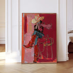 May include: Abstract art print of a person in a cowboy hat and boots, seated on a saddle. The artwork features a red and pink color palette with gold accents. The person is wearing a red shirt, blue jeans, and red boots.