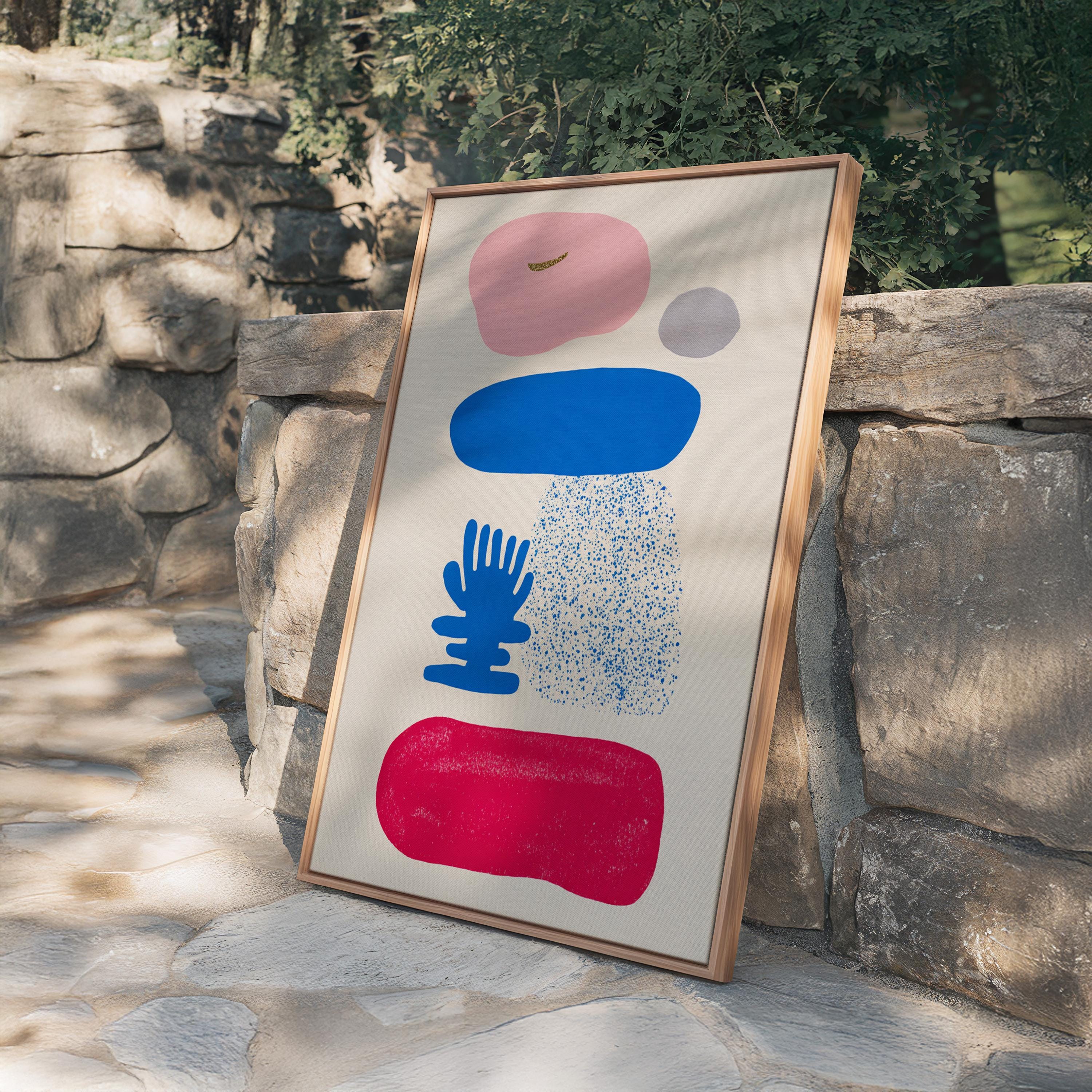 May include: Abstract art print featuring a beige background with colorful shapes. The design includes a pink, blue, red, and gray shapes. The artwork is framed in a natural wood frame, adding a touch of warmth to the modern design.