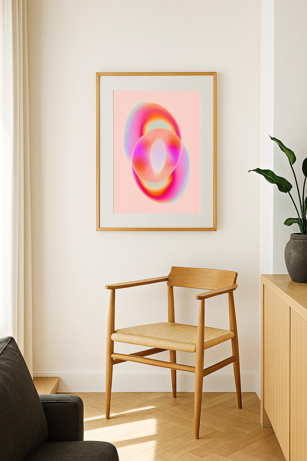 May include: A framed abstract art print featuring two overlapping, ring-like shapes. The artwork showcases a gradient of colors, including pink, orange, and blue hues. The print is displayed on a light pink background, within a gold-colored frame. Below the artwork is a wooden chair with a woven seat.