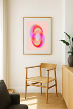 May include: A framed abstract art print featuring two overlapping, ring-like shapes. The artwork showcases a gradient of colors, including pink, orange, and blue hues. The print is displayed on a light pink background, within a gold-colored frame. Below the artwork is a wooden chair with a woven seat.