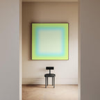 May include: A modern interior scene featuring a large square artwork with a gradient of green, blue, and white hues, framed in black. Below, a small, black chair with a circular seat and a rectangular backrest stands on a light-colored wooden floor.