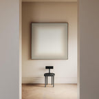 May include: A minimalist interior scene featuring a large, square artwork with a soft, gradient color scheme. Below, a small, dark gray chair with a circular seat and a rectangular backrest stands on a light-colored wooden floor.