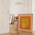 May include: A large, framed artwork with a gradient of orange and yellow hues leans against a white wall. The artwork is in a light wood frame. A wooden chair and other canvases are visible in the background, suggesting an art studio or gallery setting.
