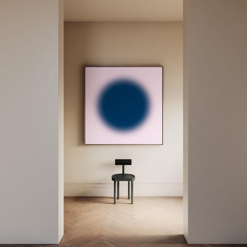 May include: A square wall art piece featuring a large, dark blue circular gradient centered on a pale pink background, displayed above a black stool with a flat, rectangular backrest and four cylindrical legs on a wooden floor.
