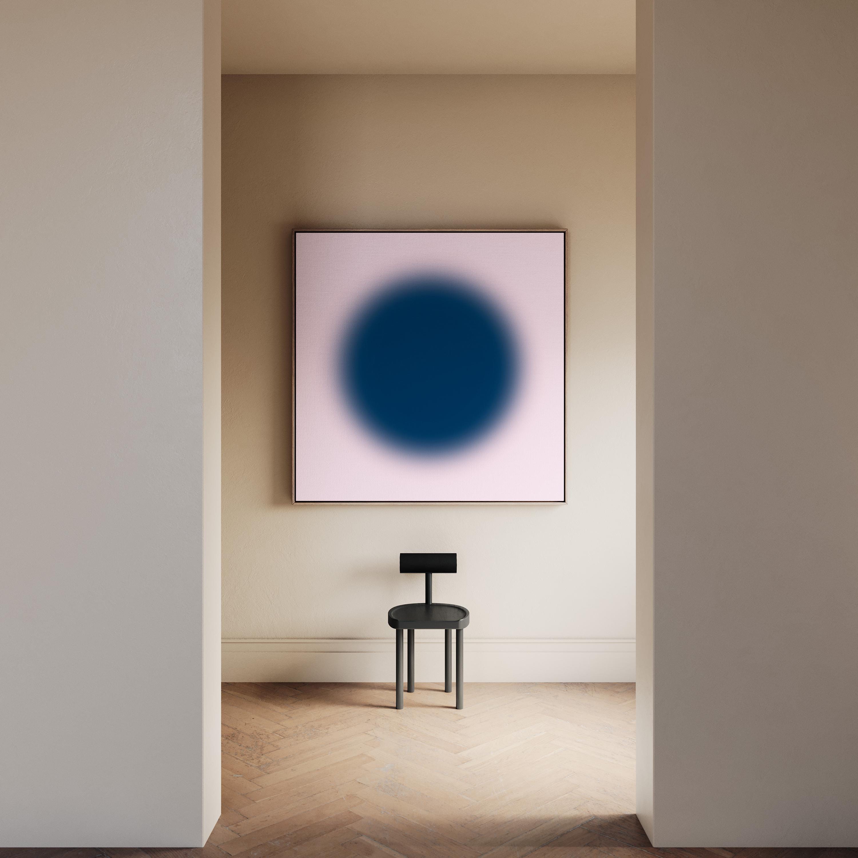 May include: A square wall art piece featuring a large, dark blue circular gradient centered on a pale pink background, displayed above a black stool with a flat, rectangular backrest and four cylindrical legs on a wooden floor.