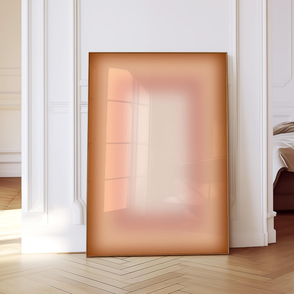 May include: A large, rectangular framed artwork leans against a white wall. The artwork features a gradient of colors, transitioning from a warm, reddish-brown at the edges to a lighter, peachy center. The frame is a thin, metallic gold color.