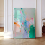 May include: Abstract art print with a gold frame. The artwork features a vibrant mix of colors, including green, blue, pink, and orange, with brushstrokes and splatters. The painting is displayed against a white wall and a light wood floor.