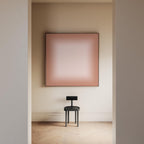 May include: A square wall art piece featuring a soft gradient from a dusty rose pink center to a darker terracotta border, framed in a thin dark edge. Below it sits a small black stool with a circular seat and a rectangular backrest.