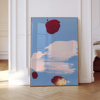 May include: Abstract art print featuring a light blue background with large brushstrokes of pink and white. Three maroon-colored shapes are scattered across the canvas. The artwork is framed in a thin, gold-colored frame.