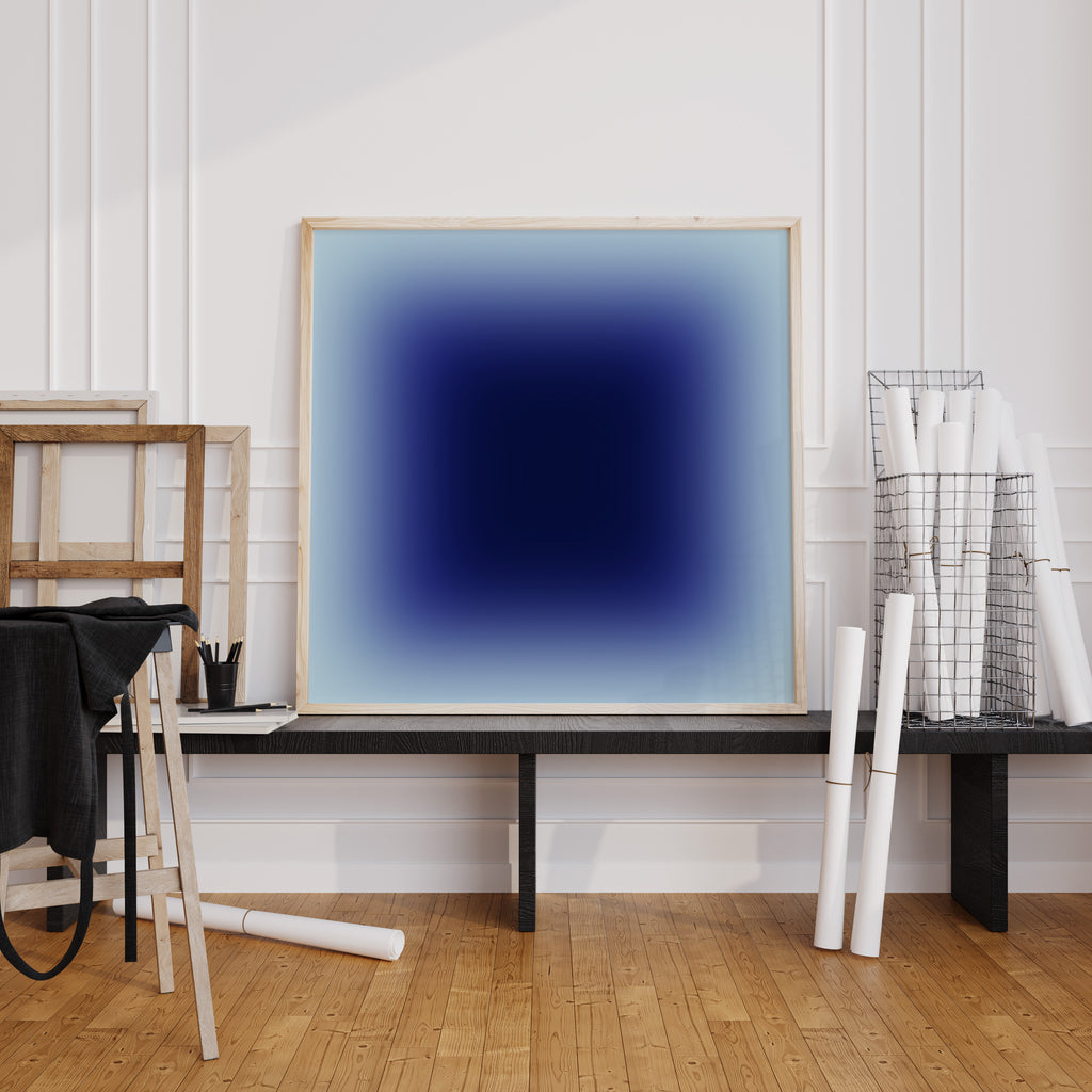 Abstract gradient artwork in indigo and sky blue tones framed in light wood, minimalist studio setting.